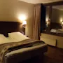 En seng med hvitt sengetøy er i et svakt opplyst hotellrom, flankert av to veggmonterte lamper. Et stort vindu med lukkede gardiner vender mot sengen.
