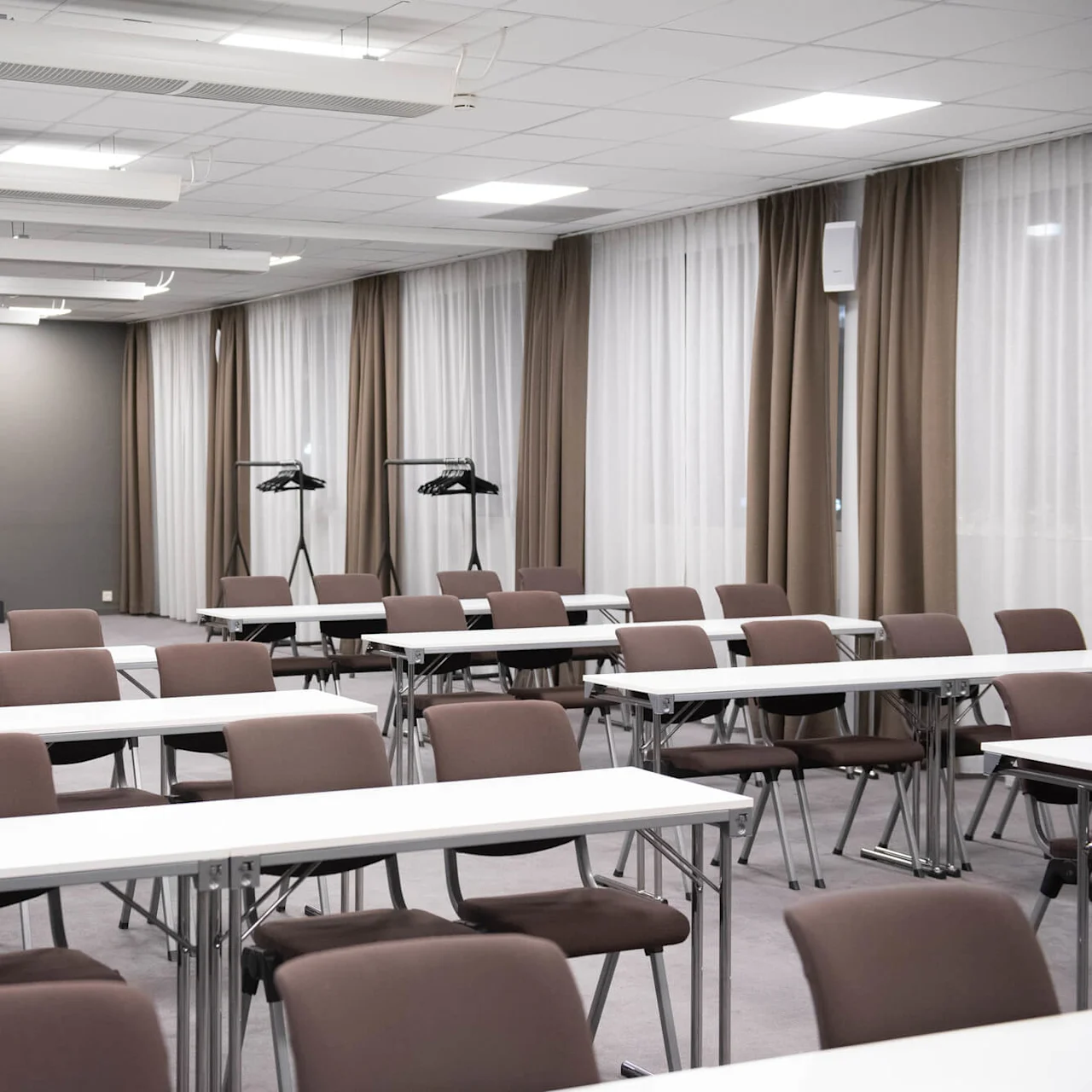 Classroom with empty brown chairs and white tables, arranged in rows. Coatracks and white curtains line the wall. A clock and sideboard with decor are at the back.