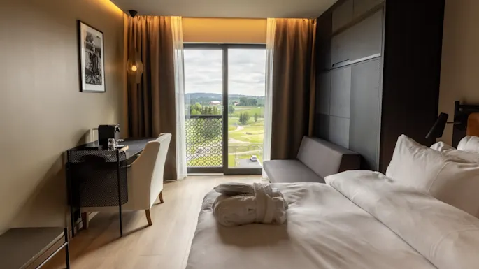 A modern hotel room with a neatly made bed, stylish furniture, and a large window revealing a scenic outdoor view.