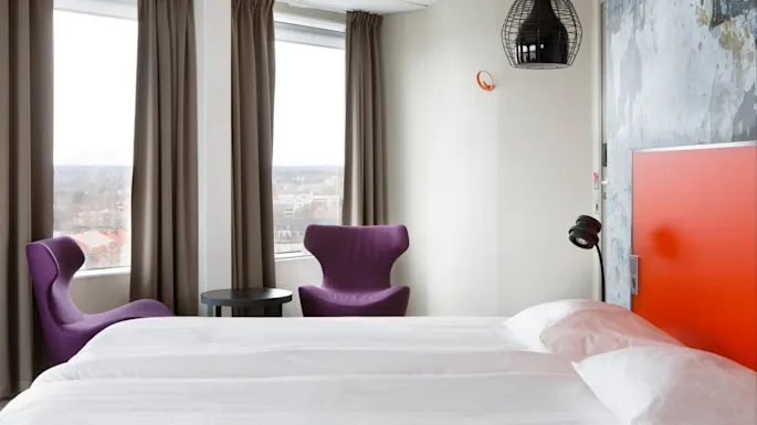 En pent oppredd seng med hvitt sengetøy står foran to lilla stoler og et rundt bord, plassert nær store vinduer med beige gardiner. En moderne pendellampe henger over.