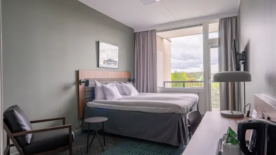 Seng er pent oppredd i et moderne hotellrom. En lenestol står ved et lite bord. Gardindekket vindu gir utsikt til utendørs. Et skrivebord har en lampe og et brett med fasiliteter.