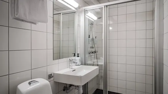 Et rent, moderne bad med hvite flislagte vegger, en vask, toalett og en glassinnkapslet dusj. Friske håndklær henger på et stativ.