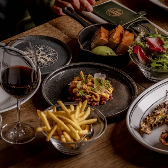 En närbild på ett restaurangbord dukat med rätter som grillad ryggbiff, råbiff, pommes frites, en grönsallad, grillat bröd och grillade havskräftor bredvid ett glas rött vin.