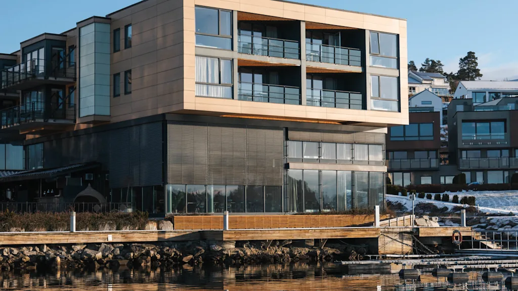 En moderne, fler-etagers bygning reflekteres i roligt vand, omgivet af andre bygninger under en klar blå himmel.
