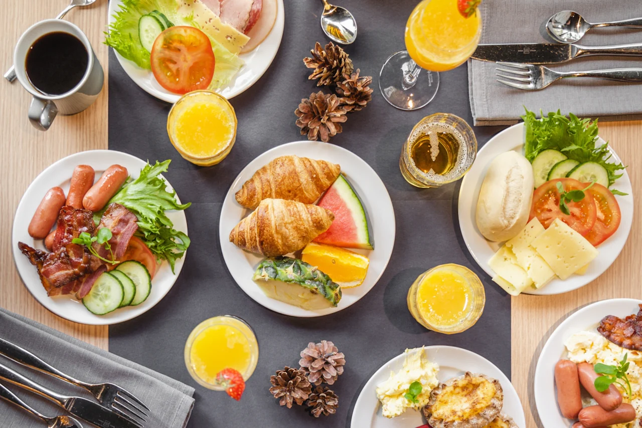 Lautasia aamiaista, mukaan lukien croissantteja, makkaroita ja juustoa, järjestetty pöydälle juomien ja käpyjen kanssa, harmaiden lautasliinojen ja ruokailuvälineiden ympäröimänä.