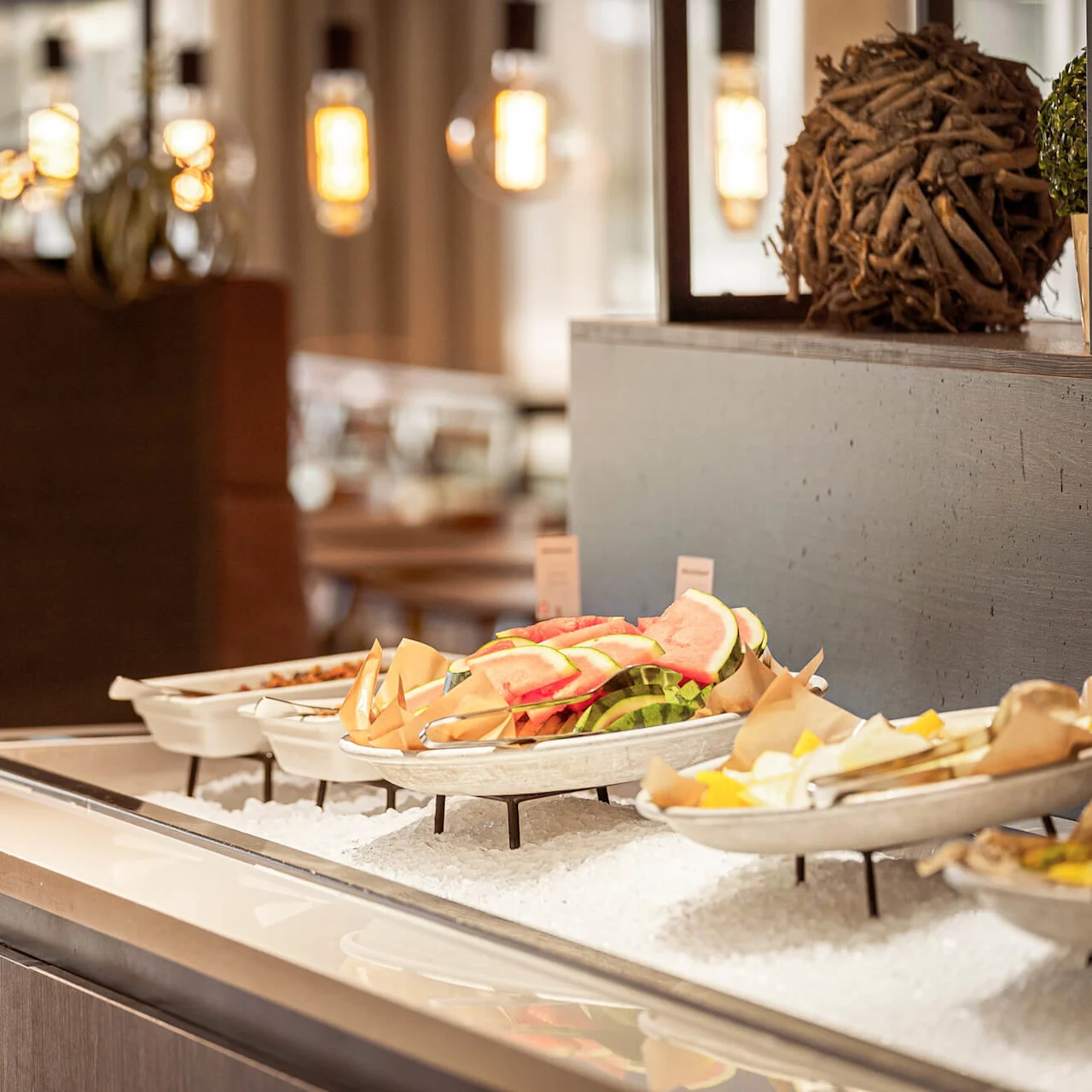Platters of fresh fruit on ice sit in a brightly lit buffet setting with modern decor elements and warm lighting.