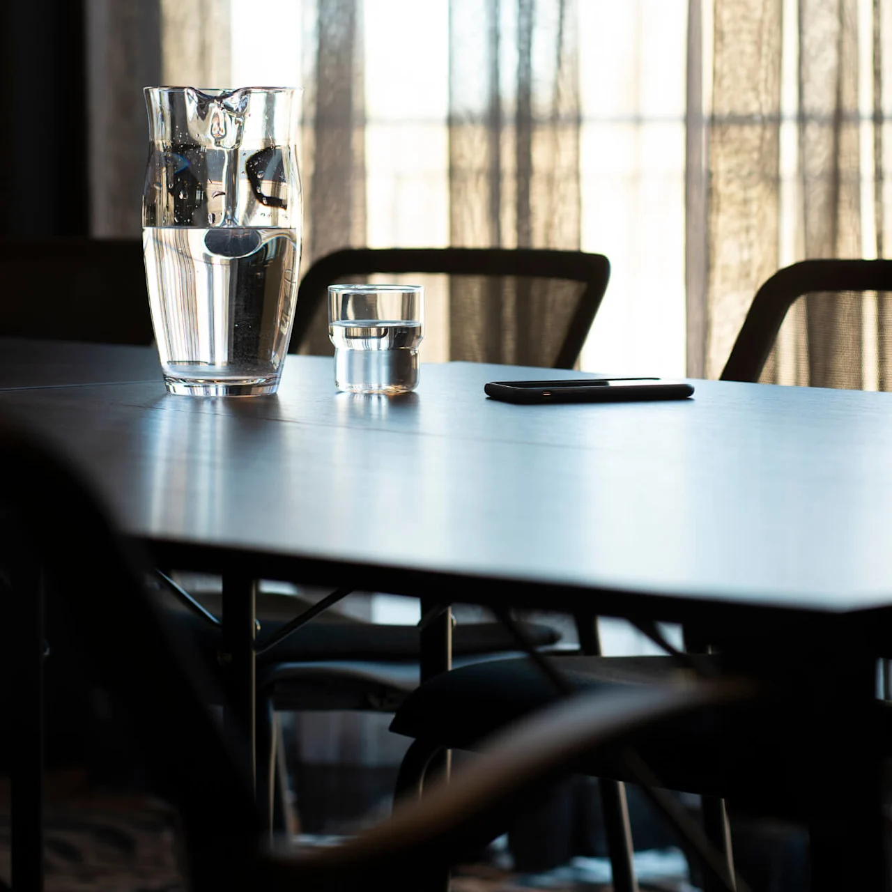 A glass pitcher and a small glass of water are on a table, accompanied by a smartphone, set against sheer curtains filtering sunlight in a meeting room.