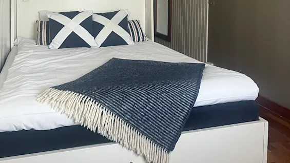 Bed with white and blue pillows and a blue fringed blanket, situated centrally in a clean, minimal bedroom with wood flooring, striped walls, and visible closet hangers in the background.