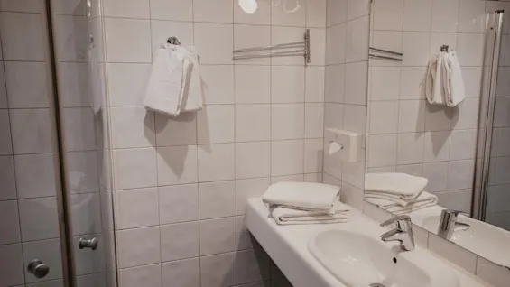 Et rent, moderne badeværelse med hvide flisevægge, en vask, spejl og bruser. Friske hvide håndklæder er pænt arrangeret.