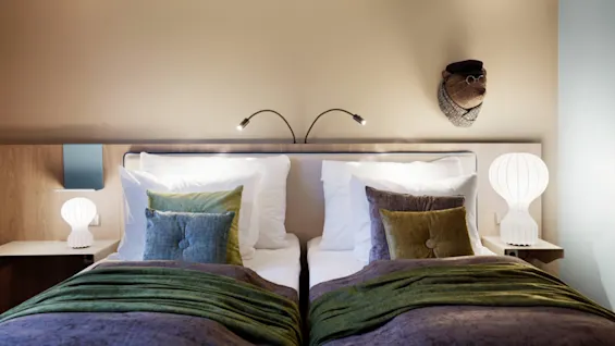 A neatly made twin bed with green and gray bedding and decorative pillows is illuminated by two reading lamps. The cozy room has white bedside lamps and a decorative mask on the wall.