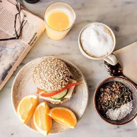En morgenmadsspredning på et marmorbord med en sandwich, appelsinskiver, kaffe, juice, og granola, ledsaget af læsebriller på en avis.