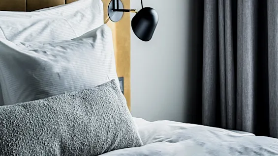 A bed with white linen and a gray textured pillow rests against a tan upholstered headboard. A black wall lamp is attached beside it, under a round ceiling light fixture.
