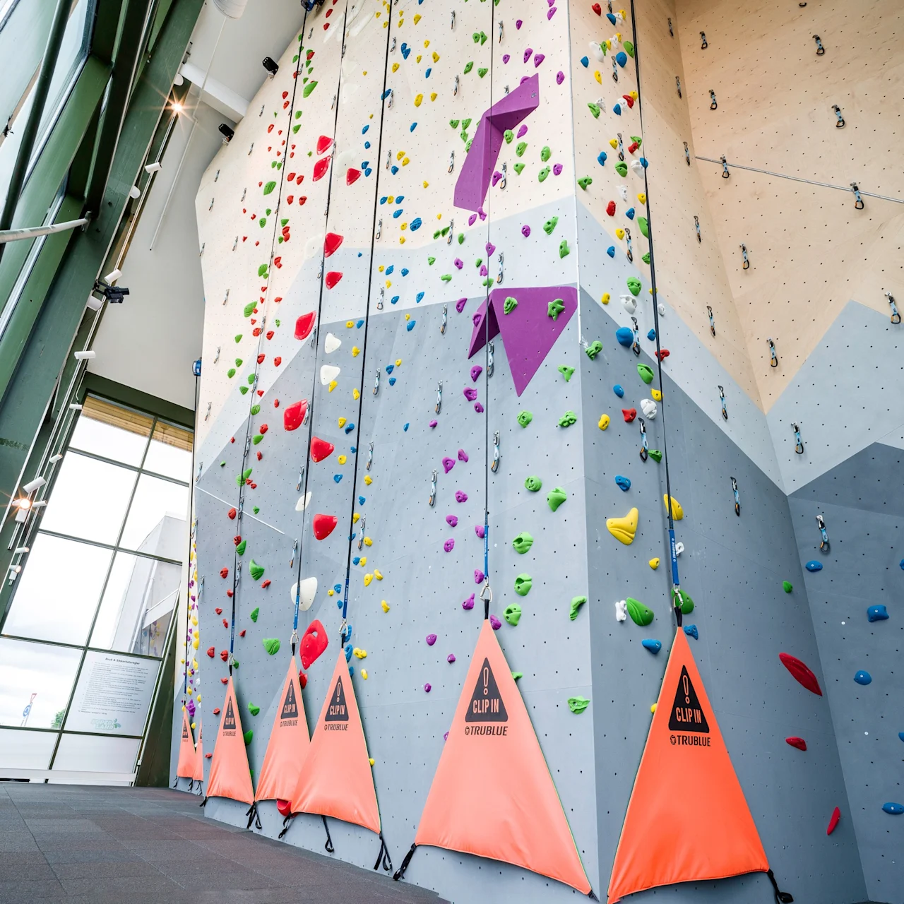 A tall indoor rock climbing wall with colorful holds and safety equipment. Climbers can scale the various routes. Situated in a spacious room with large windows. Text: "CLIP IN. TRUBLUE."