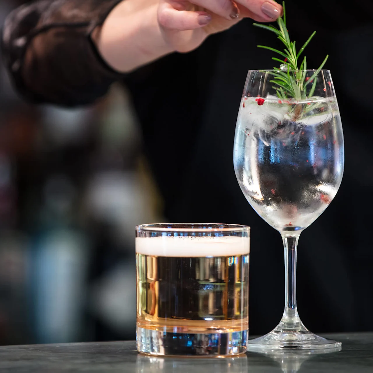 A hand garnishes a gin and tonic with a sprig of rosemary; a half-full beer glass beside it, set on a bar with a blurred background.