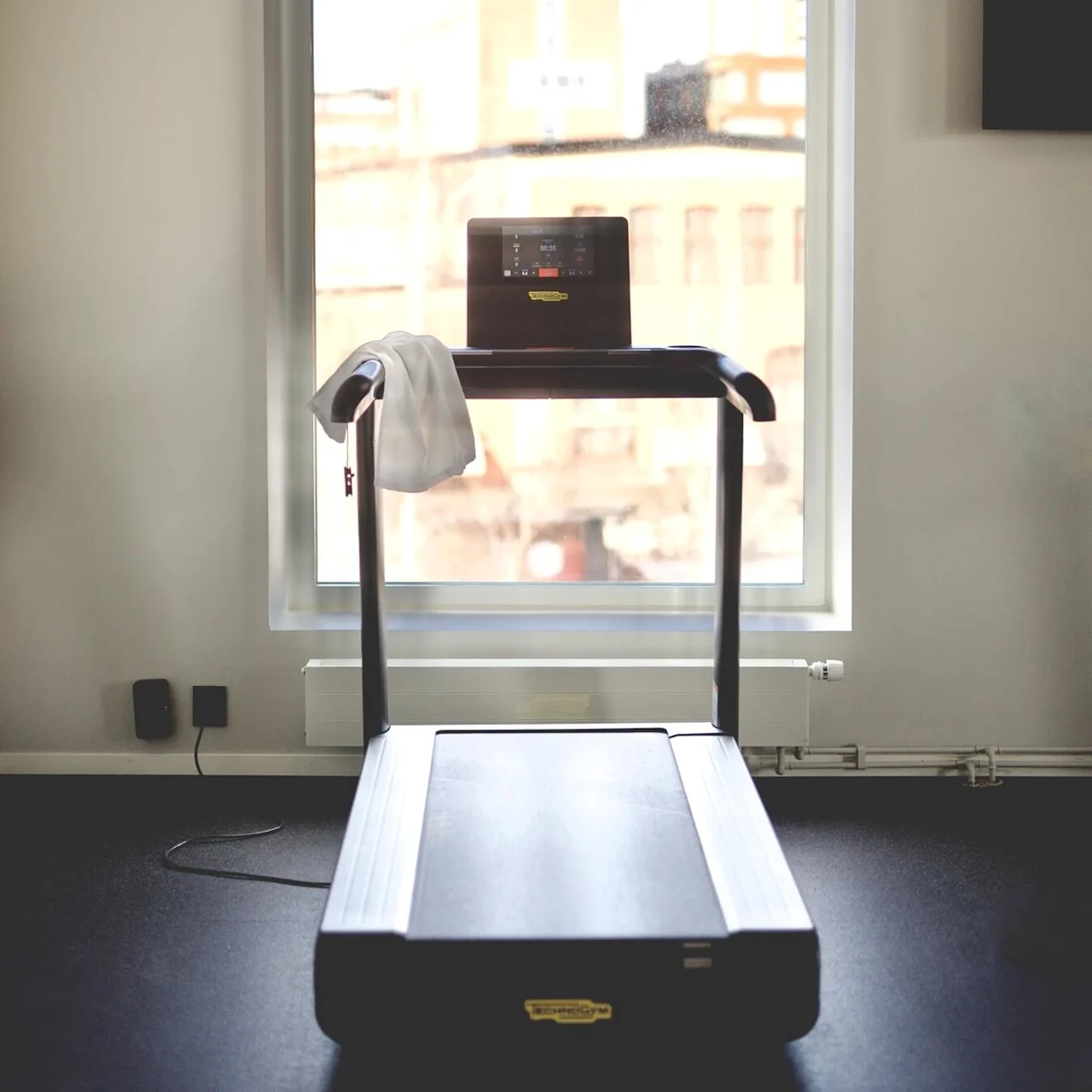 A treadmill with a towel draped over it, positioned near a window showing city buildings.