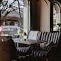 Randiga stolar omger ett litet bord med en växt och lampa i ett mysigt kafé. Stora fönster visar parkerade bilar utanför, och krukväxter pryder interiören.