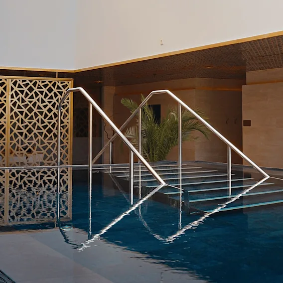 En inomhuspool på ett spa med mörkblått vatten och metallräcken som leder ner för trappan. En gyllene geometrisk skärm och en palm dekorerar bakgrunden.