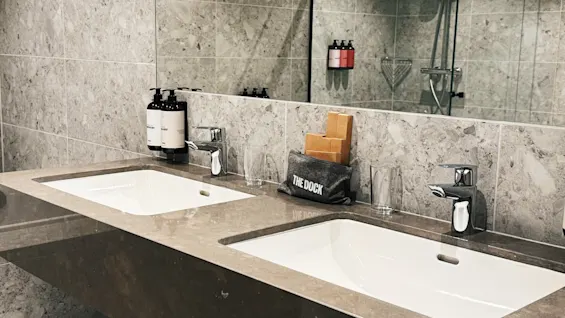 Ett elegant, modernt badrum med dubbla handfat, marmorklädda väggar och en dusch synlig i spegeln. Bekvämligheter inkluderar tvål och en väska märkt THE DOCK.