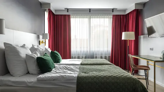 Ett mysigt hotellrum med en välbäddad dubbelsäng med mjuka gröna och vita kuddar. Varm belysning, röda gardiner och ett skrivbord vid fönstret skapar en inbjudande och avkopplande atmosfär. En TV är monterad på väggen.