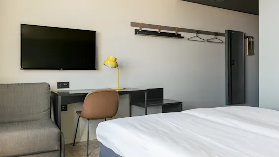 En väggmonterad TV är ovanför ett skrivbord med en gul lampa och en brun stol. Bredvid sängen syns en öppen garderob med galgar. Rummet har en modern stil.