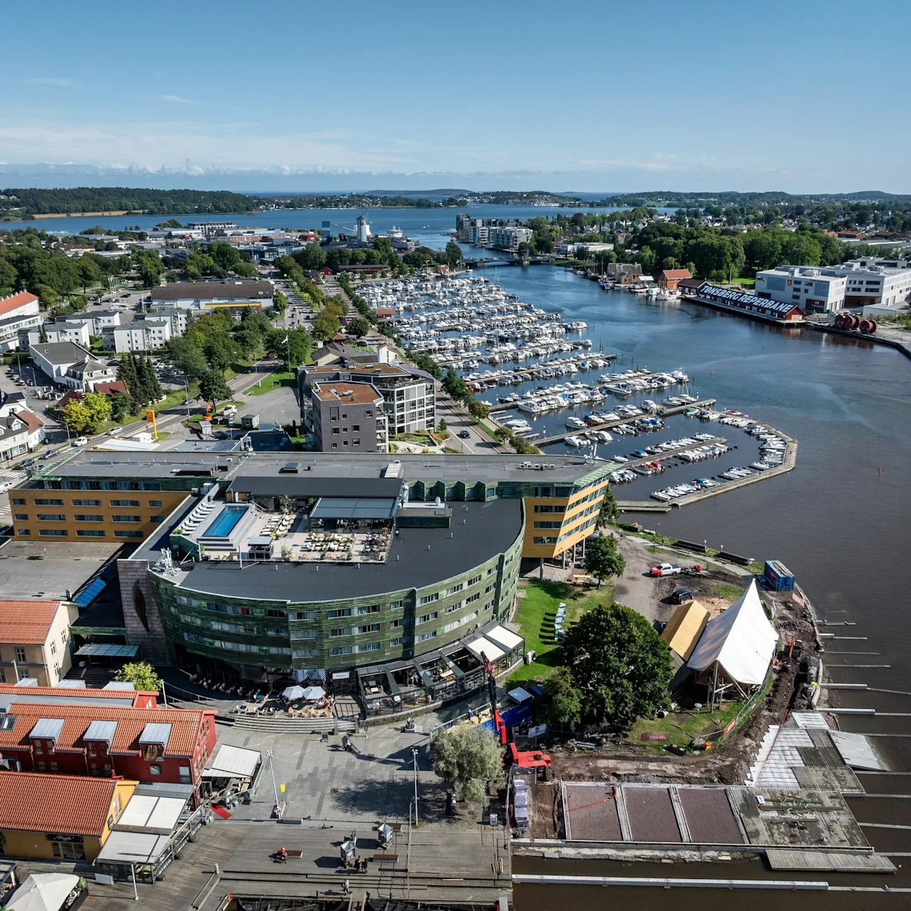 Ett böjt, modernt byggnad med gröna och gula paneler blickar ut över en småbåtshamn full med förtöjda båtar. Omkring finns bostadshus och ett färgglatt, grönt landskap mot horisonten.