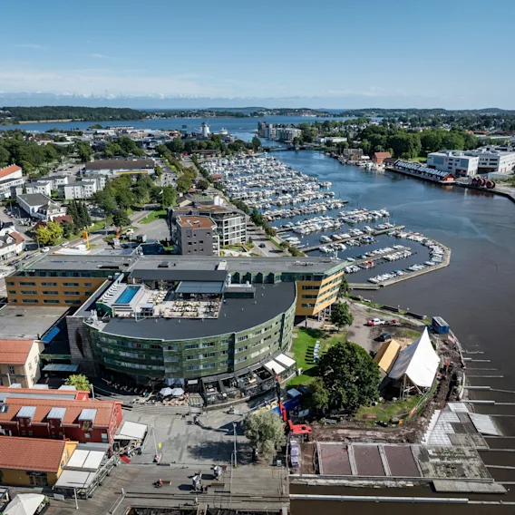En buet, moderne bygning med grønne og gule paneler ser ut over en marina full av fortøyde båter. Omkringliggende er boliger og et frodig, grønt landskap som strekker seg til horisonten.