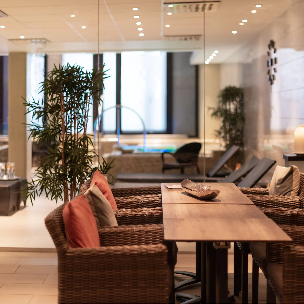 A wooden table with wicker chairs and cushions stands in a cozy spa-like room. Glass walls reveal a pool area, greenery, and soft lighting for relaxation. Text: "OTIUM".