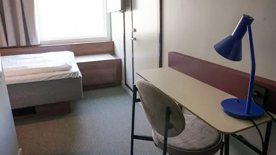 Et lite rom med en seng, et enkelt skrivebord og en blå lampe ved siden av en stol. Et lyst vindu er delvis dekket av gardiner som kaster lys på teppet.