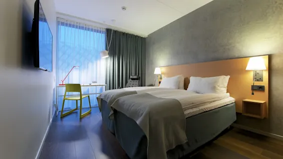 A neatly made bed with white linens stands against a concrete wall. Beside it, a desk with a chair and lamp occupies the well-lit room. A television is mounted on the opposite wall.