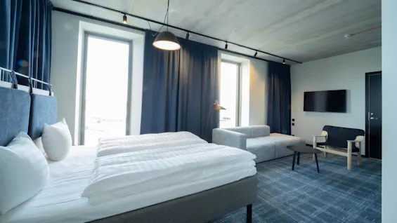 Moderni hotellihuone, jossa suuri sänky valkoisine liinavaatteineen, harmaa sohva, musta tuoli ja seinälle asennettu televisio. Siniset verhot peittävät korkeat ikkunat, lisäten tilan tuntua.