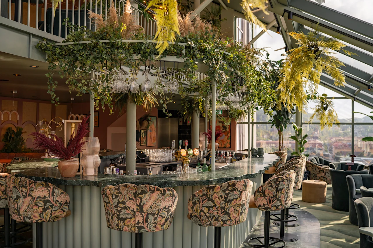 Tyylikäs baarialue, jossa on vihreä tiski, kukkakuvioiset baarituolit ja reheviä riippuvia kasveja, luoden kutsuvan ja eloisan tunnelman.