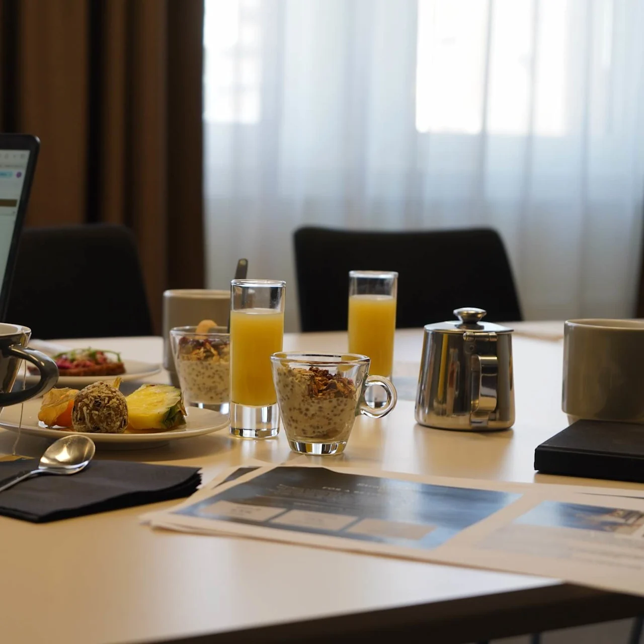 Laptop displaying a spreadsheet on a table; coffee cup, juice glasses, chia pudding, and pastries surround it in a meeting room setting. Papers and a silver teapot are nearby.