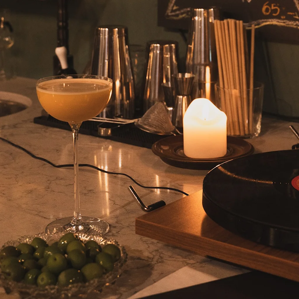 En hyggelig barscene med en cocktail, oliven, et tændt lys og en pladespiller, der skaber en afslappet atmosfære.