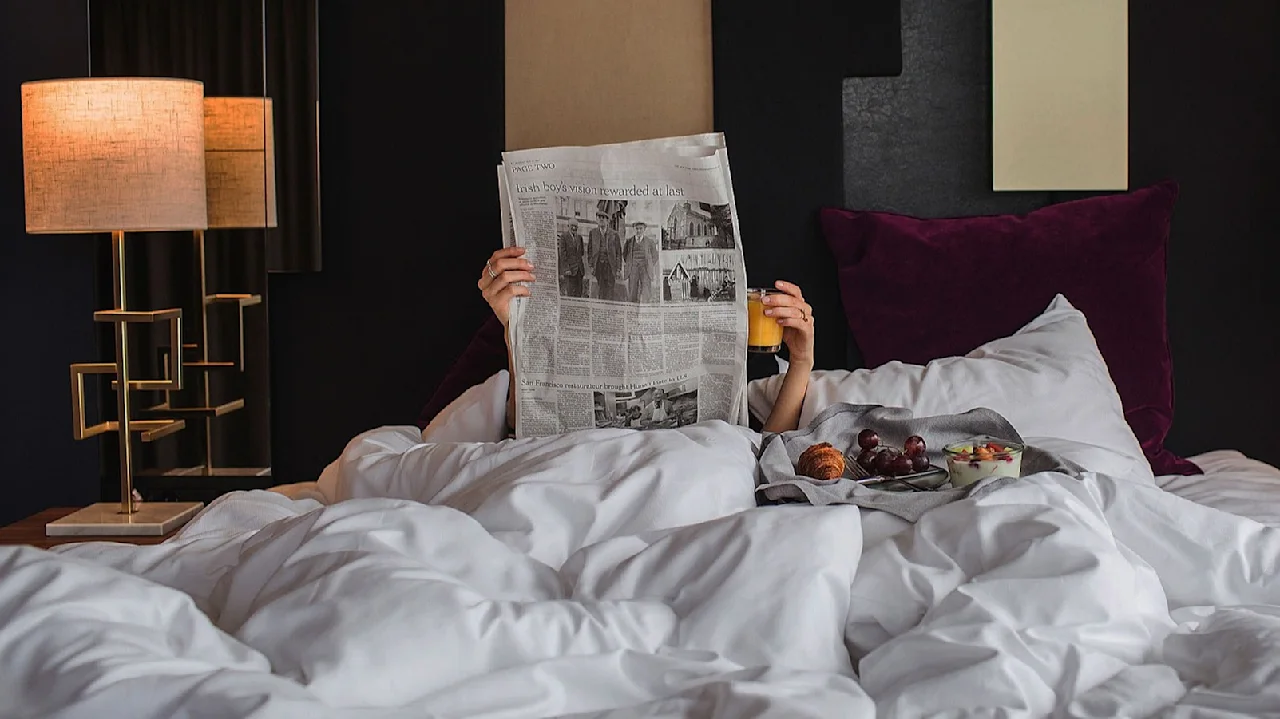 Person som läser en tidning i sängen, håller ett glas apelsinjuice; omgiven av mjuka kuddar och svagt upplyst med två sänglampor i en mysig sovrumsmiljö.