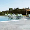 Uima-allas selkeällä sinisellä vedellä, joka heijastaa riviä tyhjiä vihreitä aurinkotuoleja. Ympärillä kivilaattoja, vehreää kasvillisuutta ja metsämaisema.