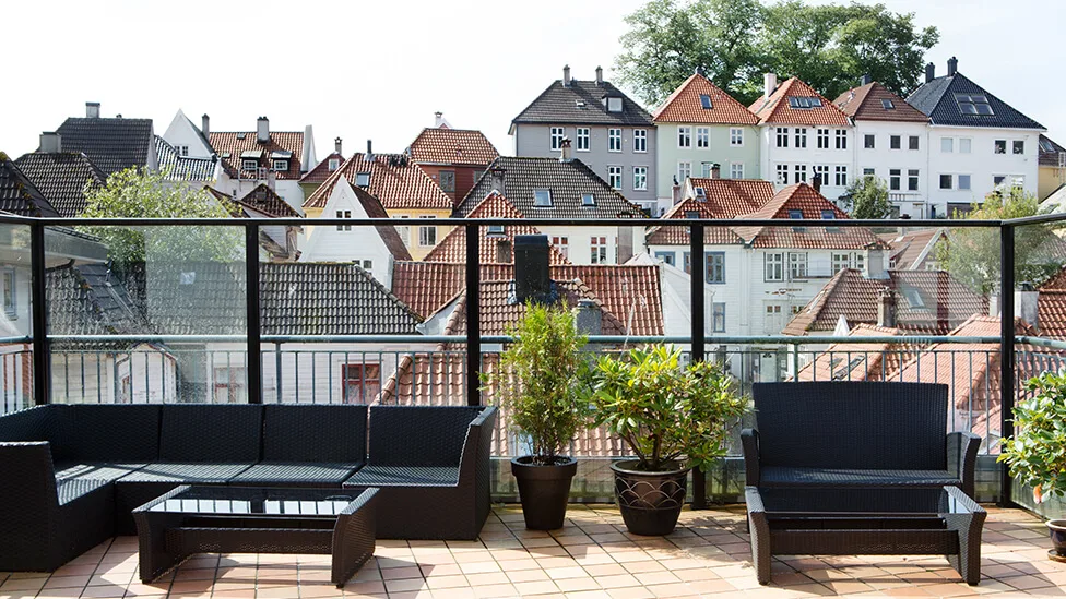 Outdoor seating arrangement features black wicker furniture and potted plants on a tiled terrace, overlooking a scenic view of charming, traditional houses with varied rooftop designs.