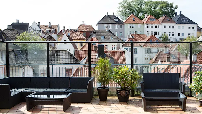 Outdoor seating arrangement features black wicker furniture and potted plants on a tiled terrace, overlooking a scenic view of charming, traditional houses with varied rooftop designs.