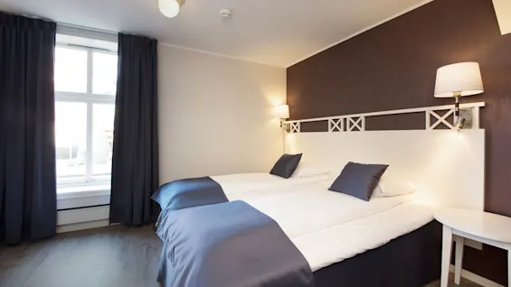 Ett välstädat sovrum med enkelsängar, blå och vit bäddning, mörka gardiner och en sänghuvudgavel i trä för en modern inredning.