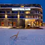 En kjelke står på snøen foran en fleretasje bygning med opplyste vinduer, navngitt "QUALITY HOTEL SKIFER." Det omkringliggende lyset skaper en koselig vinterkveldstemning.