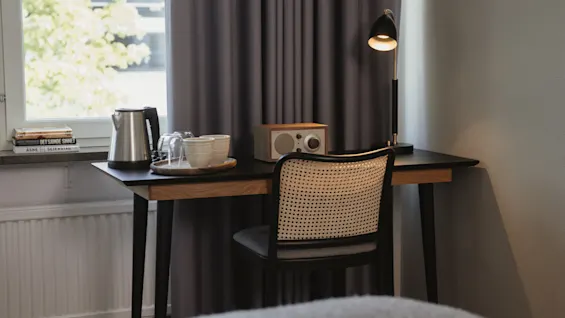 Skrivebord med en stol under, viser en lampe, radio og bakke med kedel og kopper. Plassert mot et gardin, med et vindu på siden.