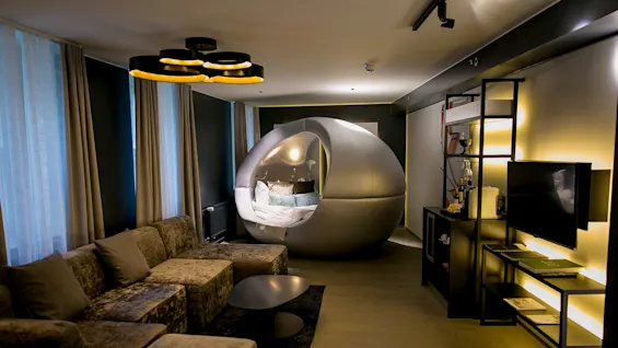 Futuristinen, pallomainen vuodepod med seinällä himmeästi valaistussa, modernissa olohuoneessa, jossa on samettinen sohvaryhmä, kuvioidut verhot ja hyllyt koriste-esineineen.