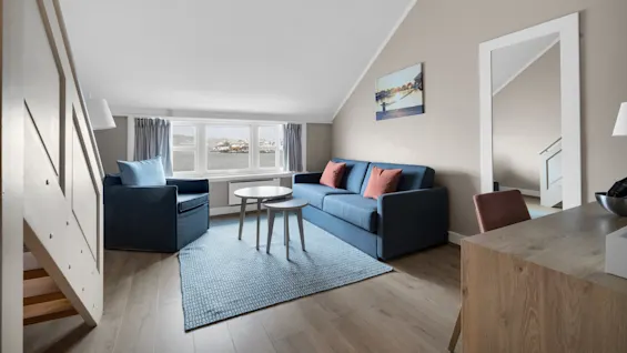 En blå sofa og lenestol står på et teppe i en lys loftsstue med store vinduer og utsikt over vannet, samt et speil på veggen.