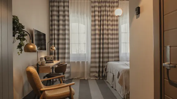 A cozy hotel room with a bed, a desk area, and a comfortable chair, featuring striped curtains and warm lighting.