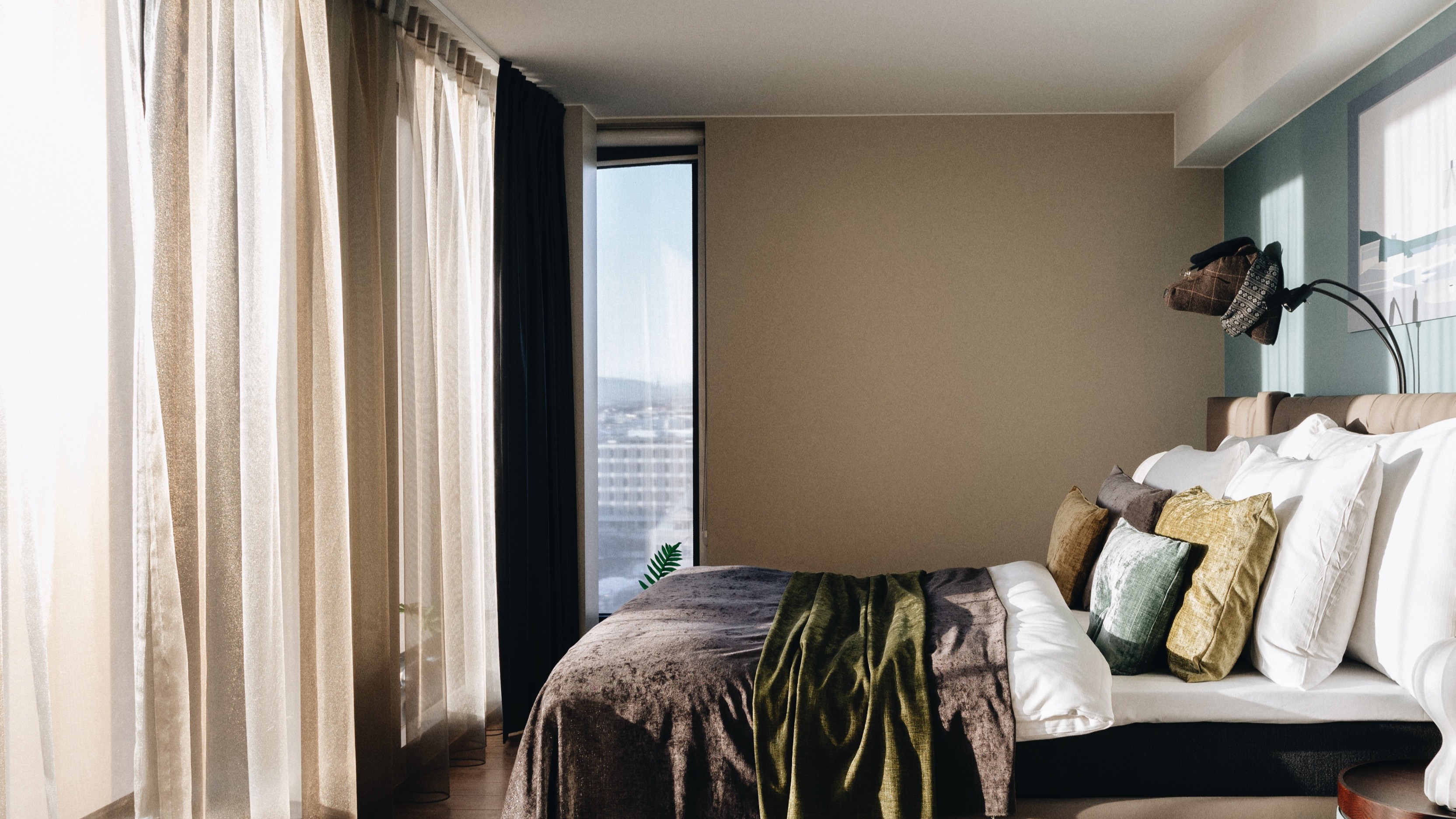 Et hyggeligt hotelværelse med stor seng, bløde puder og varmt tæppe, badet i naturligt lys fra stort vindue med udsigt over by.