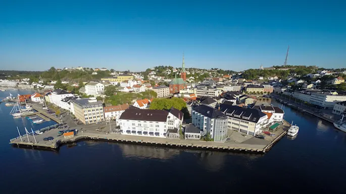 Coastal town buildings overlook a calm harbor with several boats docked; a tall church spire rises amidst green hills under a clear blue sky. 