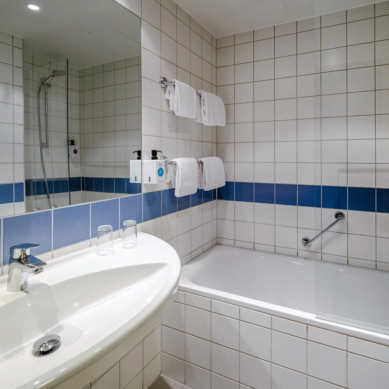 Handfat med kran och två glas, bredvid ett badkar med duschhuvud och tvåldispensrar, i ett kaklat badrum med en blå accentlinje och prydligt hängda vita handdukar.