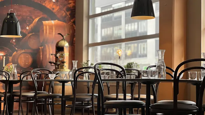 Bord med glasservise under sorte pendellamper, omgitt av stoler. Bakgrunn med et vintønnemaleri, store vinduer og planter, skaper en koselig restaurantstemning.