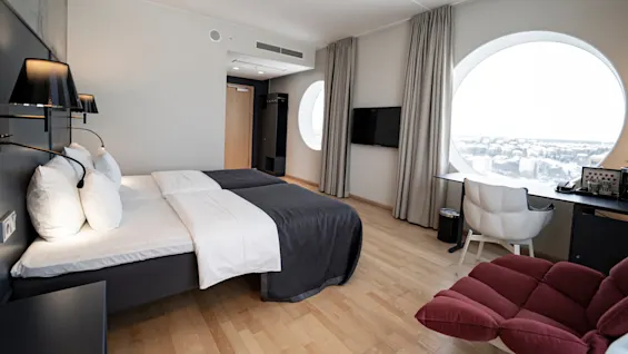 A neatly made double bed under wall lamps, in a modern hotel room. Large round window provides a cityscape view. Nearby, there’s a desk, chair, and television.