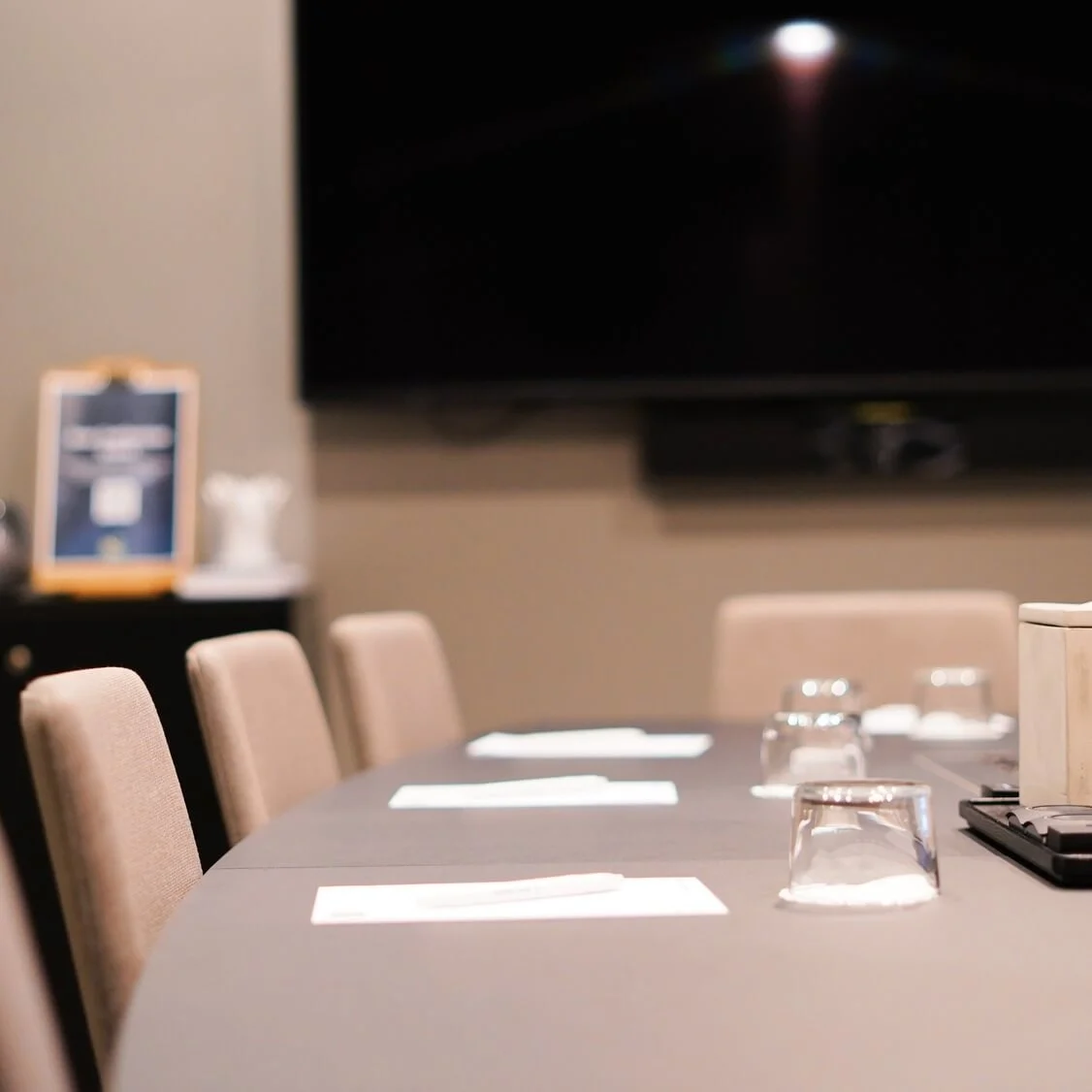 A conference table set with notepads, pens, and upside-down glasses; a decorative box in the center. A clipboard and flowers are in the background under a wall-mounted TV.