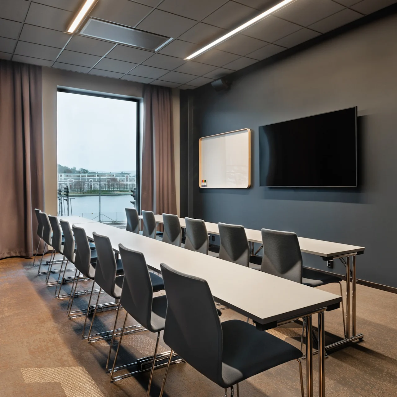 Konferanserom med rader av tomme stoler og bord vendt mot en stor skjerm og whiteboard, vindusutsikt over en bygning ved elvebredden. Nøytrale farger dominerer den moderne, minimalistiske designen.